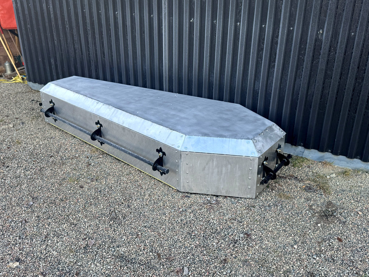 Specialbyggd takbox i aluminium formad som en gammal likkista, på grus mot svart plåtvägg