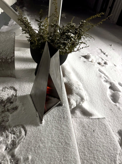 Triangel-ljuslykta tänd i snön en vinterkväll bredvid en kruka – här i blankt, obehandlat stål