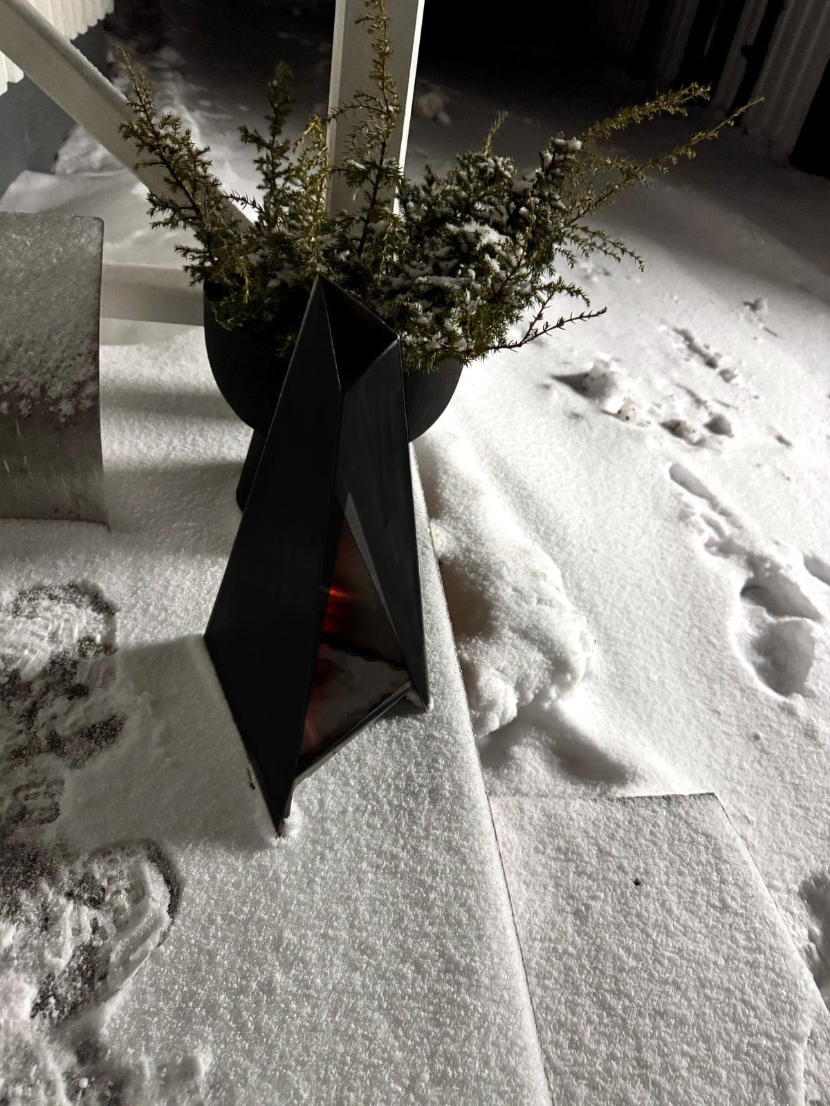 Svart Triangel-lykta i snön bredvid en kruka vid entrén
